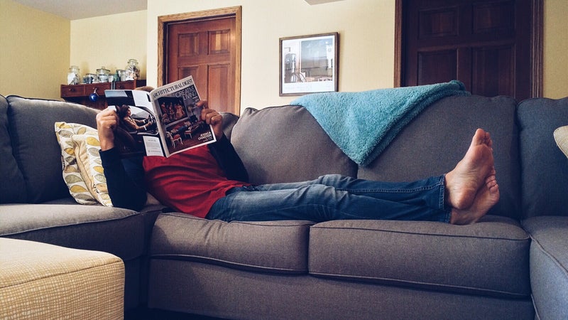 A person lounging on a gray couch reading a magazine