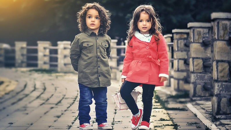 Two children standing outside on a stone walkway wearing coats